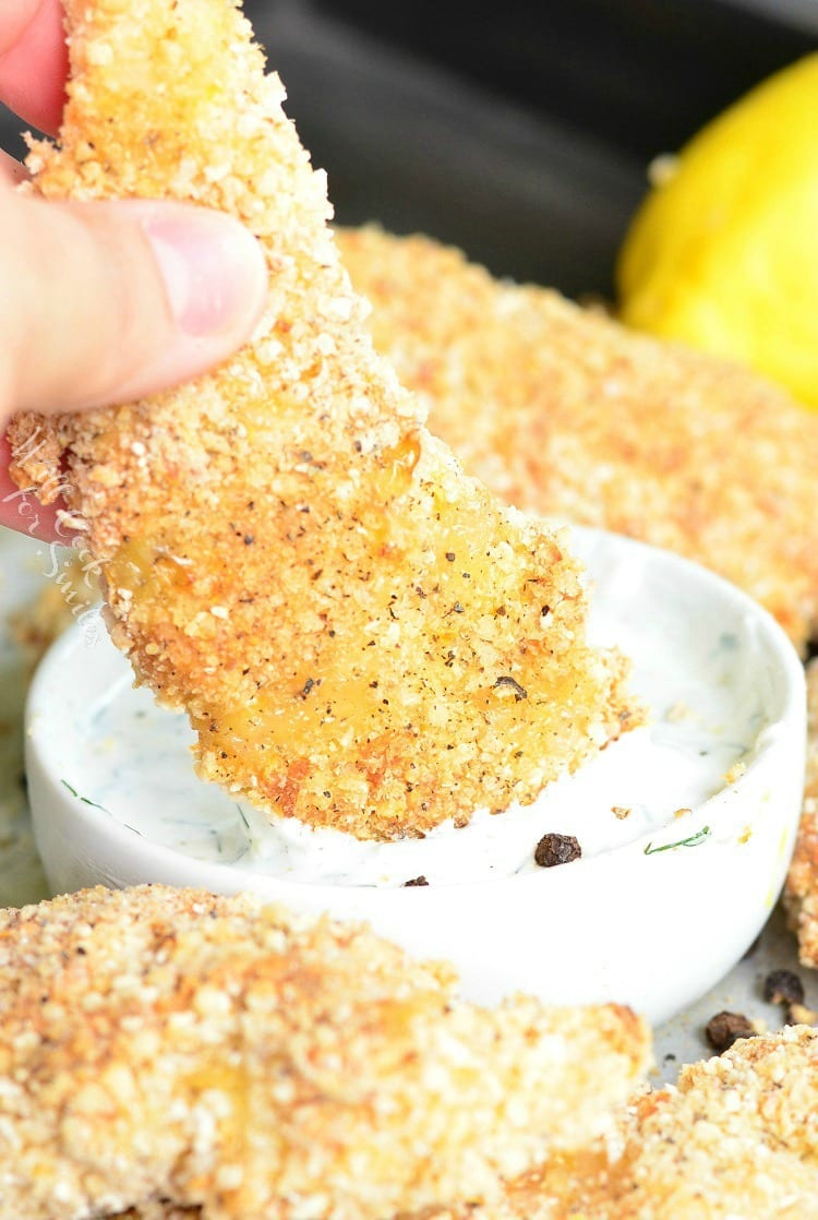 Lemon pepper baked chicken tenders around a dish of ranch with a whole lemon in the background and 1 tender being dipped into sauce.