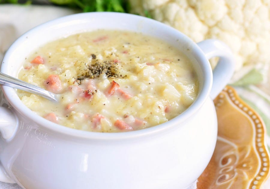 horizontal view of cauliflower sou with ham pieces visible and a metal spoon in a white bowl, on a brown plate