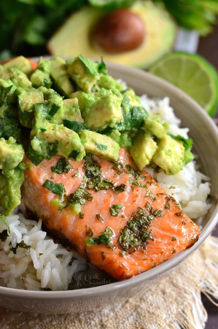 salmon topped with avocado over rice in a bowl on a table with avocado in the background  