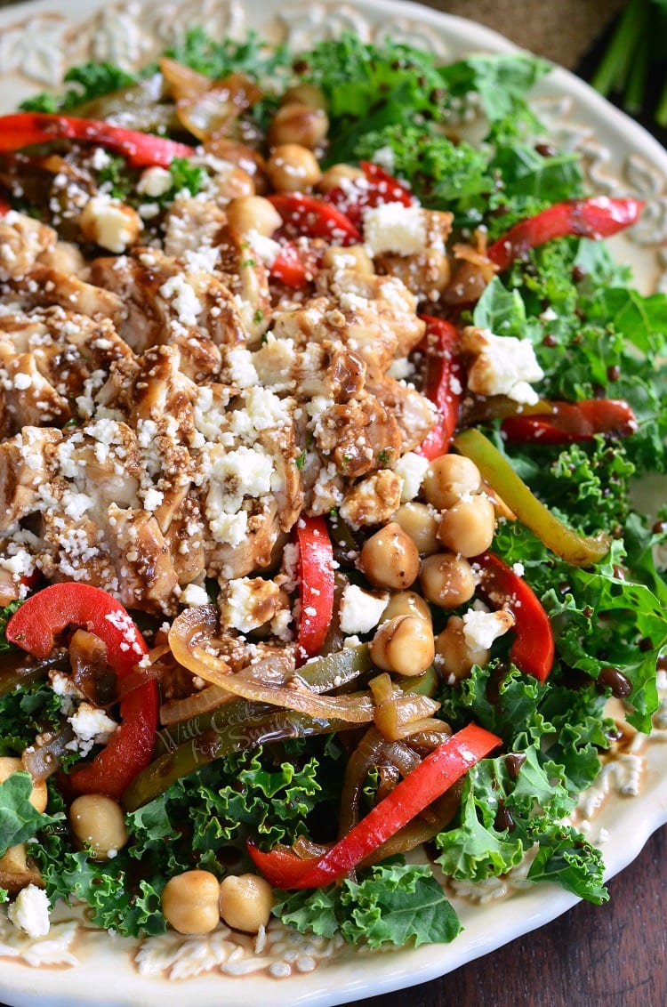 Top view of photo of kale salad topped with chicken and veggies that have been sauteed in balsamic on a white plate with dressing, chick peas, and finally, lots of feta cheese.