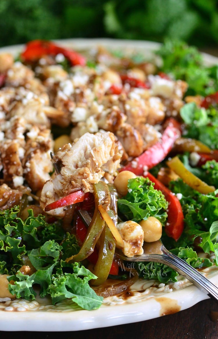 kale salad topped with chicken and veggies that have been sauteed in balsamic on a white plate with a fork dressing, chick peas, and finally, lots of feta cheese.