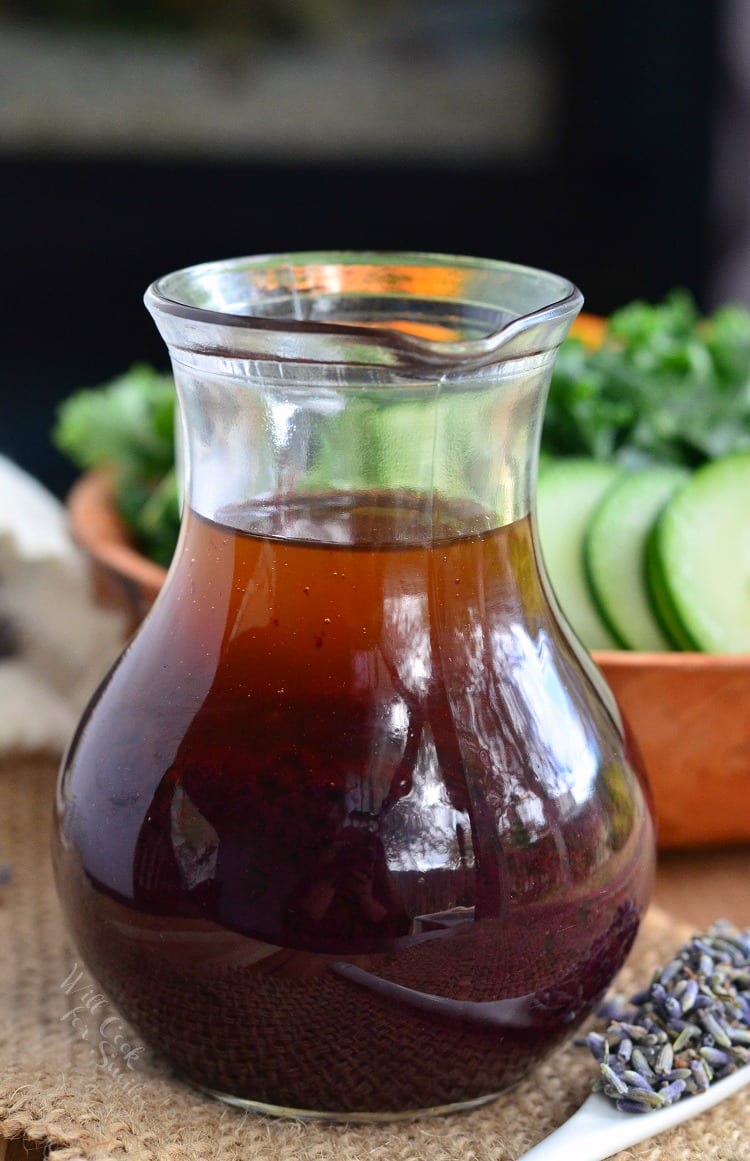 Blueberry Lavender Vinaigrette in a glass container on the right is some lavender on a white spoon in the backroom is a salad in a brown bowl 