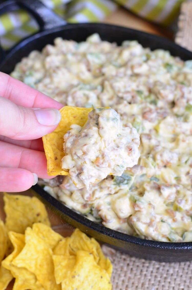 dipping a tortilla chip into Cheesy Sausage Peppers and Onions Dip that is in a cast iron skillet 