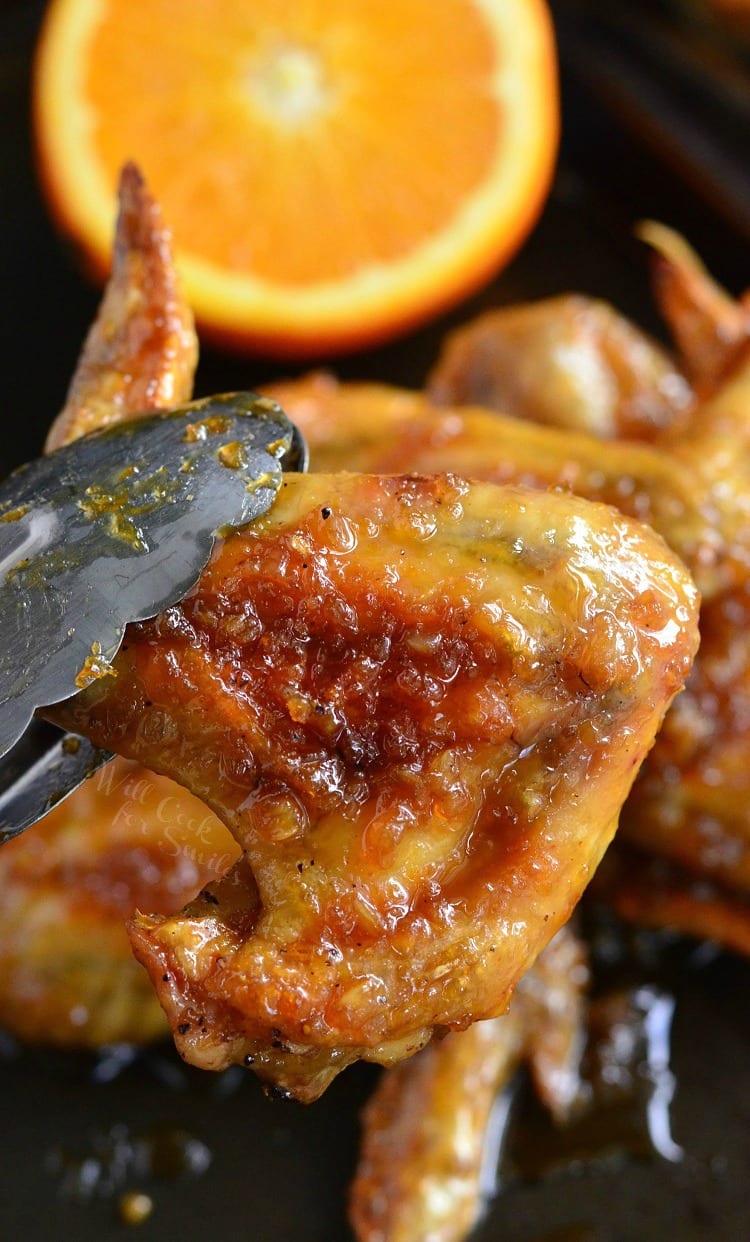 lifting Spicy Orange Chicken Wing with metal tongs off of baking sheet with a orange in the background 