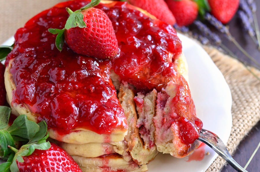 Horizonal photo of Strawberry Lavender Pancakes with strawberries and strawberries syrup on top on a white plate with a fork 