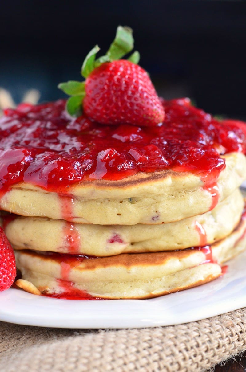 stack of strawberry pancakes with strawberry sauce on top and a strawberry in the middle on a white plate.