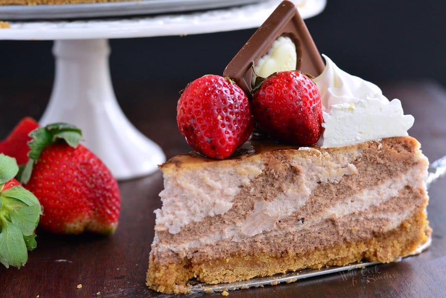 Chocolate Strawberry Marbled Cheesecake on a table with strawberries around it and a cake stand in the background 