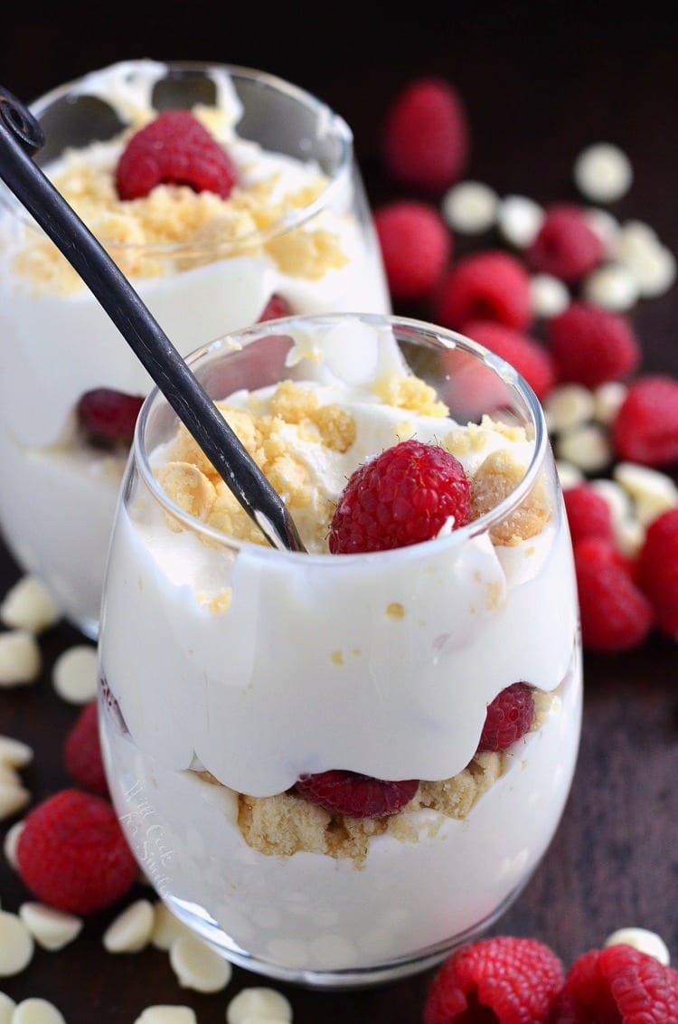 Raspberry White Chocolate Mousse Parfait in a stemless wine glass with a spoon on a table with white chocolate and raspberries around it 