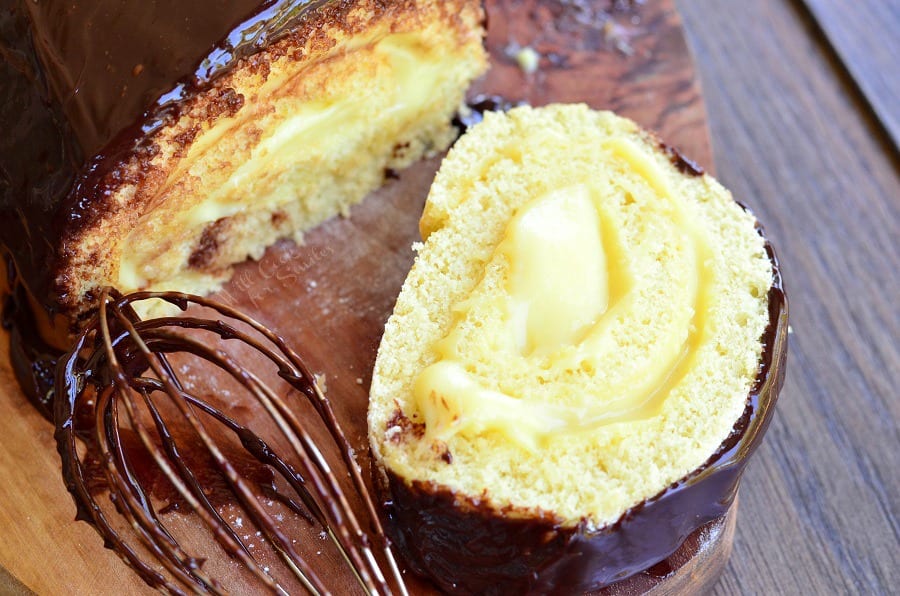 Boston Cream Cake Roll with a slice on a wood cutting board with a whisk with chocolate on it 