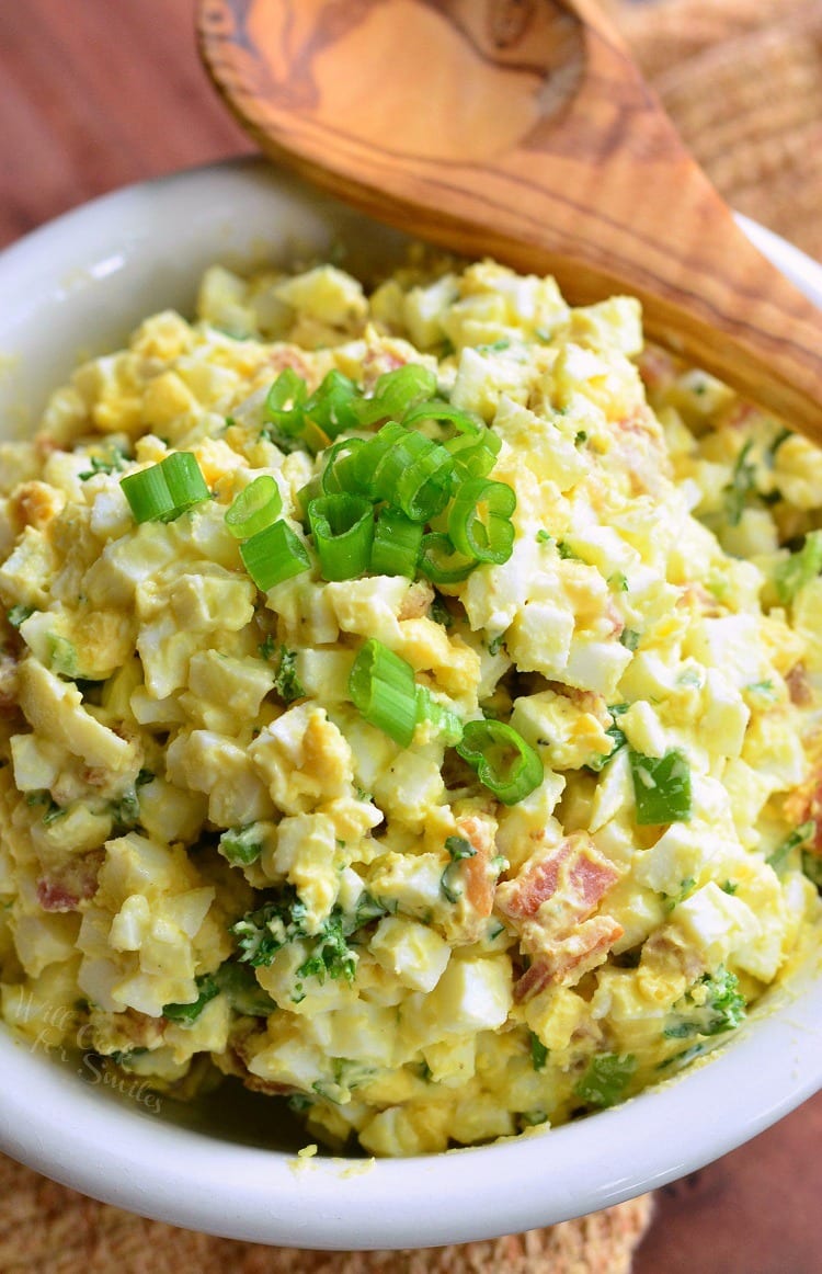 top view image of egg salad in a white bowl with green onions as garnish and a wooden spoon across the top 
