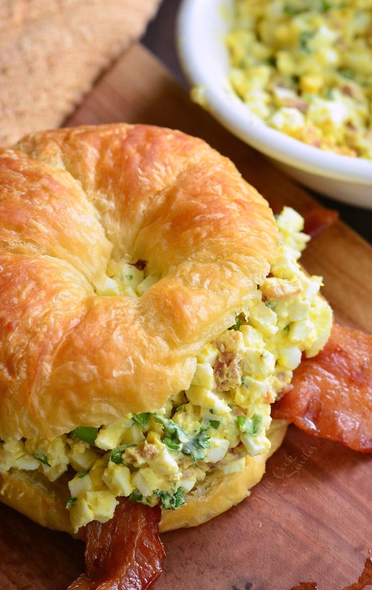 top view of egg salad  sandwich with bacon on a cutting board with bowl of egg salad in the background 