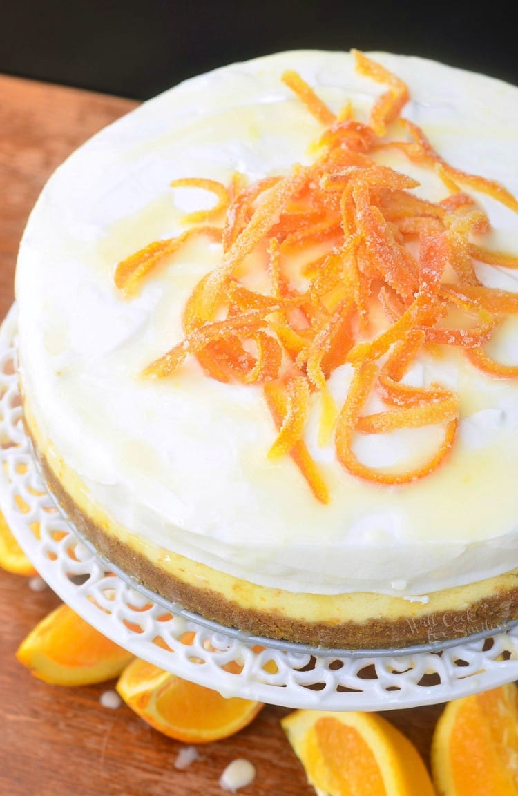 top view Orange Creamsicle Cheesecake on a cake stand with candy oranges on top and orange slices around the bottom of cake stand on a table 