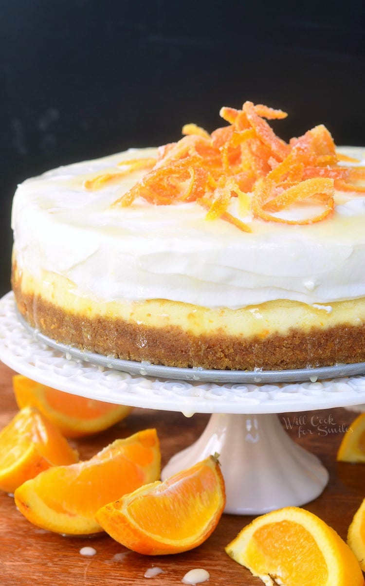 Orange Creamsicle Cheesecake on a cake stand with candy oranges on top and orange slices around the bottom of cake stand on a table 