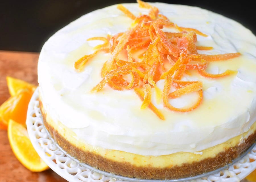 Orange Creamsicle Cheesecake on a cake stand with candy oranges on top and orange slices around the bottom of cake stand on a table 