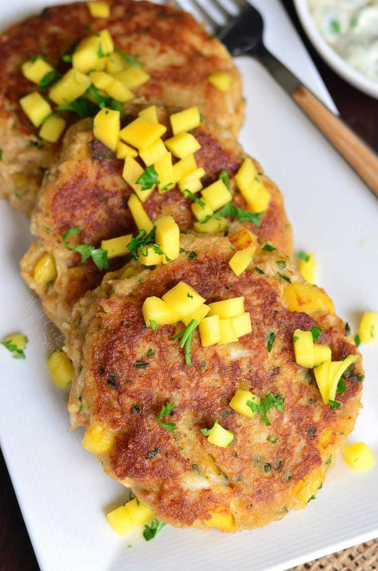 four large mango crab cakes side by site on a white plate with more mango on top.