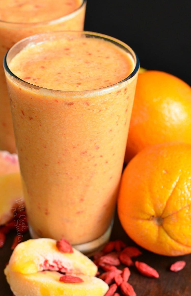 2 glasses of Creamy Peach and Goji Berry Smoothie in a glass on a table with a orange, peach, and goji berry around it