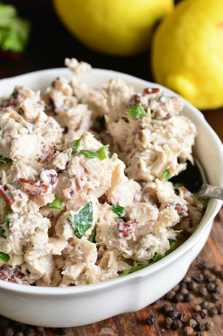 Lemon Pepper Rotisserie Chicken Salad in a white bowl on a wood table with a lemon in the background and peppercorns to the right in font of the bowl 