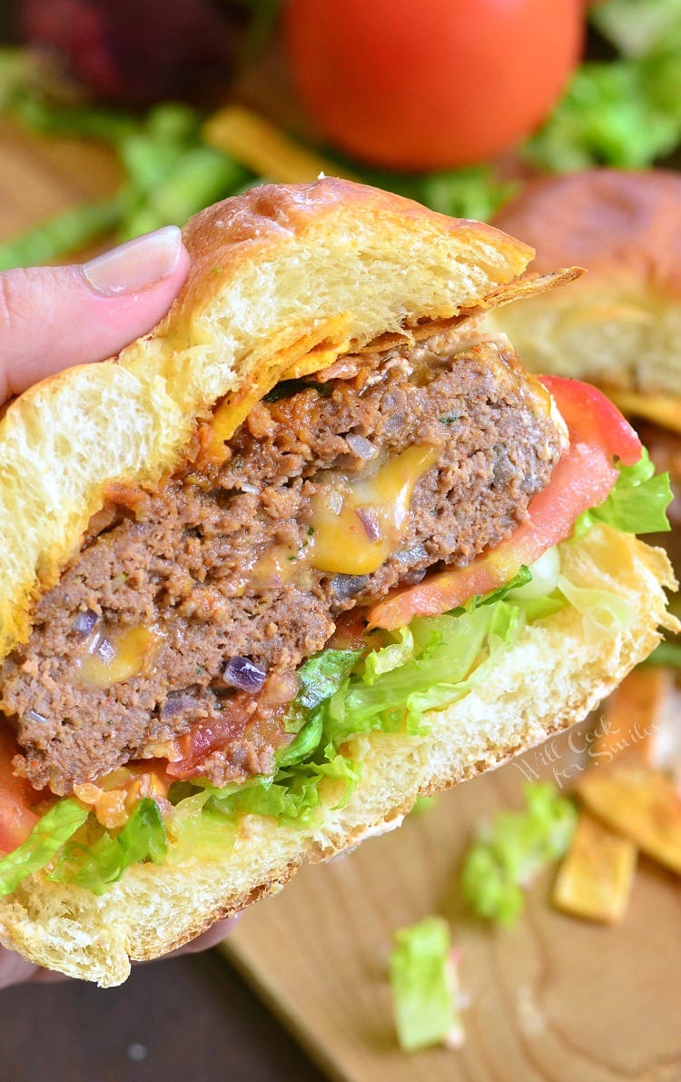 holding Stuffed Taco Burger on a bun cut in half with lettuce and tomato with tomato in the background 