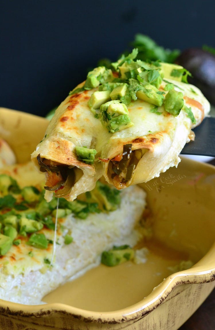 lifting Vegetable Enchiladas in a yellow backing dish with diced avocado and cilantro on top out with a spatula 