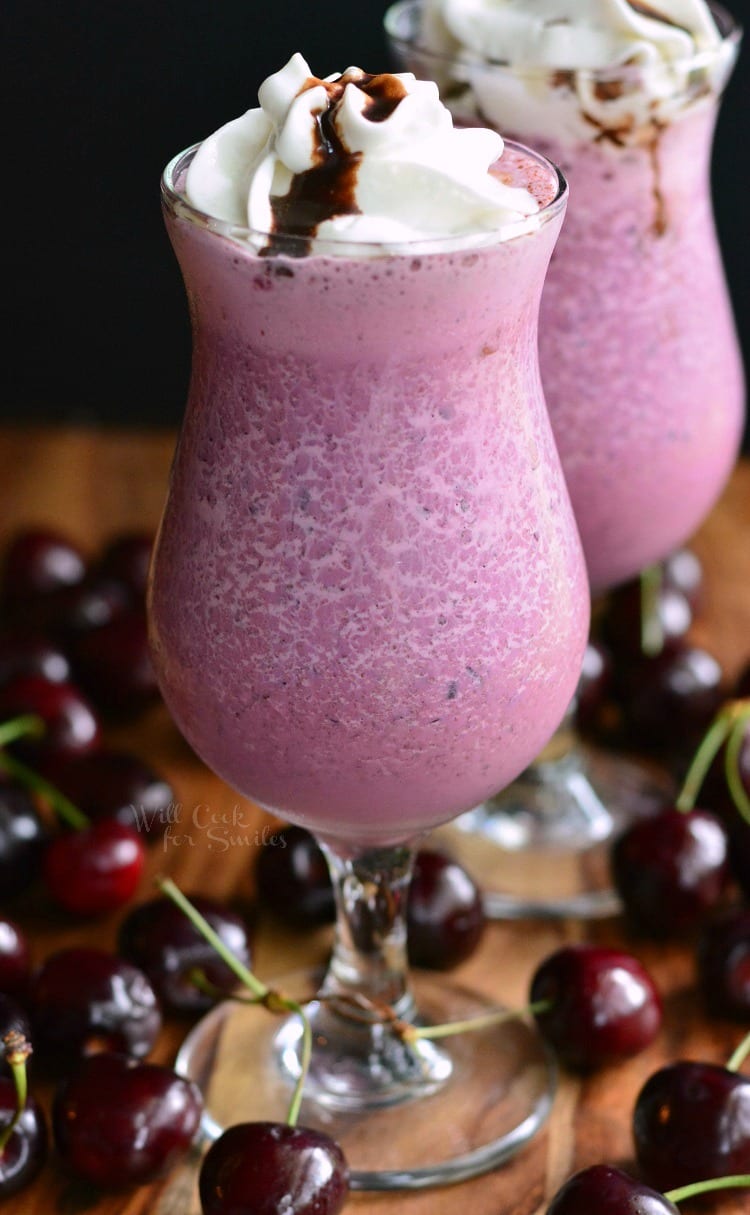 2 Chocolate Covered Cherry Milkshake in a glass with whip cream and cholate drizzled on top on a wood table with cherries around it