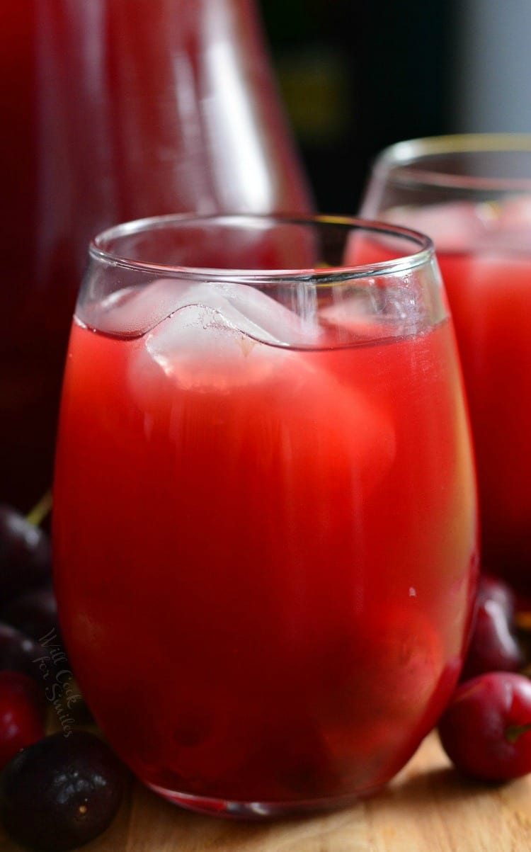 Homemade Cherry Arnold Palmer in a stemless wine glass on a table with a pitcher in the background and cherries around it 