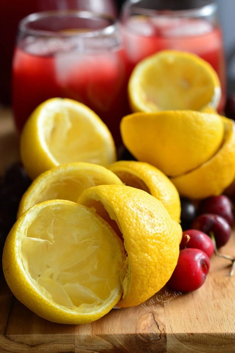 Squeezed lemons and cherries in a cutting board and Cherry Arnold Palmer in glassed in the background all on a cutting board 