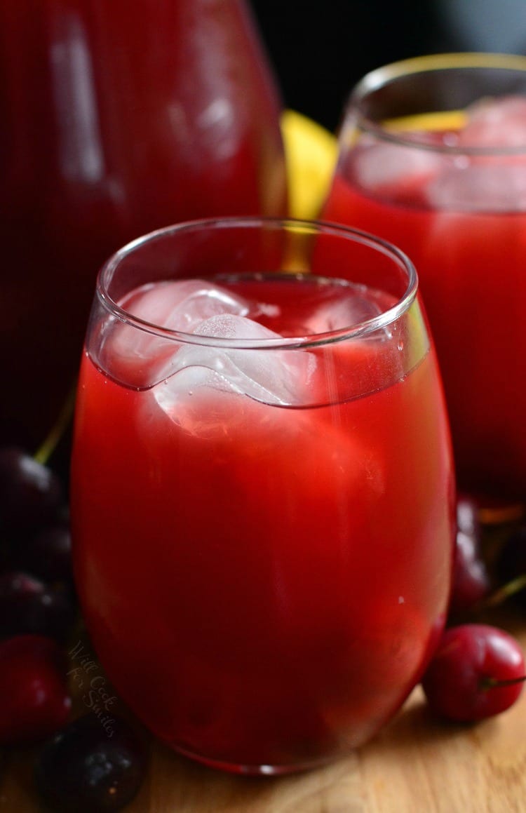 a pitcher and 2 glasses of Cherry Arnold Palmer on a cutting board with cherries around it 