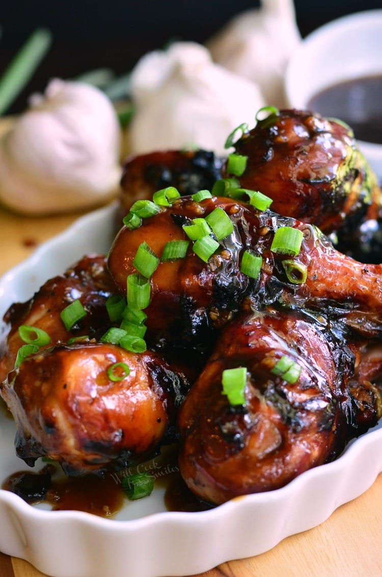 Sweet Garlic Grilled Chicken Drumsticks with green onions on top on a serving plate with garlic in the background