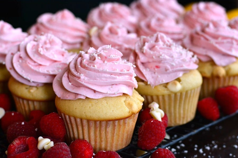 horizonal photo of cupcakes with pink frosting on top and white chocolate chips and raspberries around it.