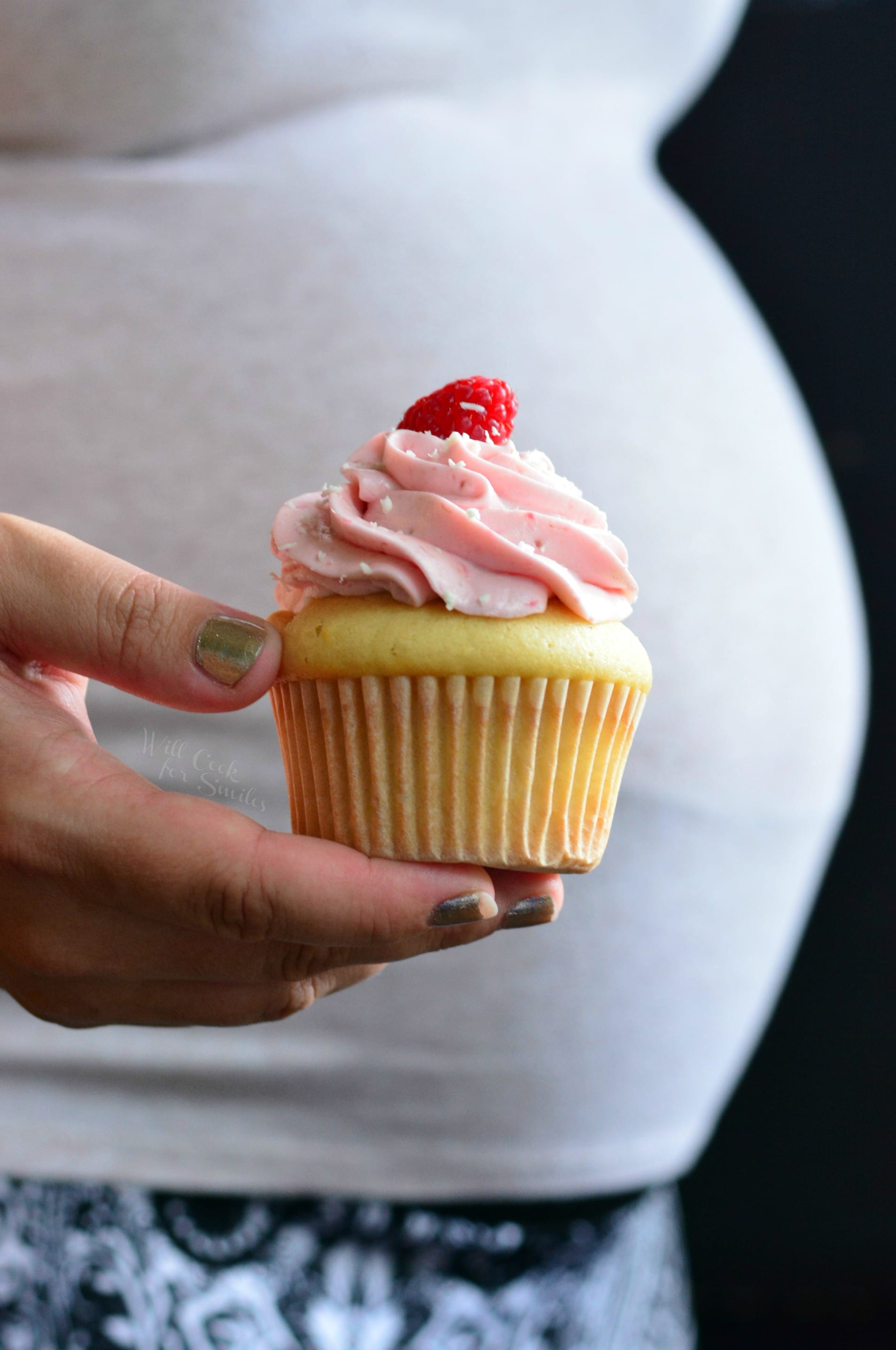 holding a cupcake with pink frosting and a raspberry on top in front of a pregnant belly.
