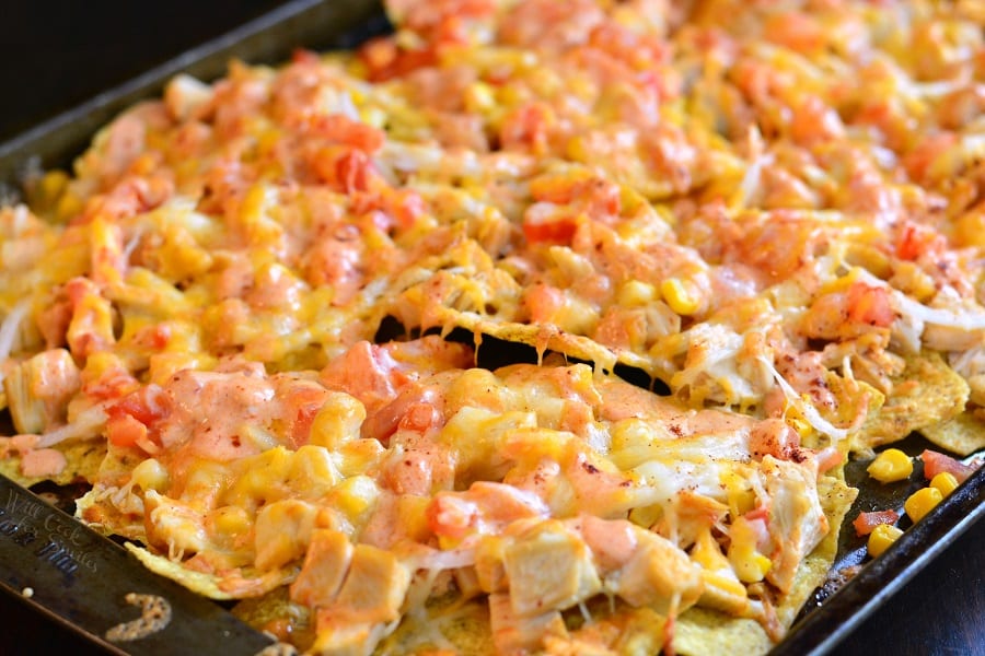 A close-up of a tray filled with loaded nachos topped with melted cheese, diced tomatoes, corn, and chunks of chicken. The nachos are golden and crispy, with toppings evenly spread across the surface.