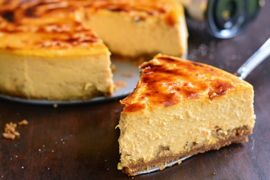 slice of Creme Brulee Pumpkin Cheesecake on a pie spatula on a wood table with the rest of the cheesecake in the background