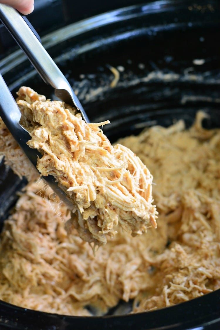 shredded chicken in a crockpot with some being picked up with tongs. 
