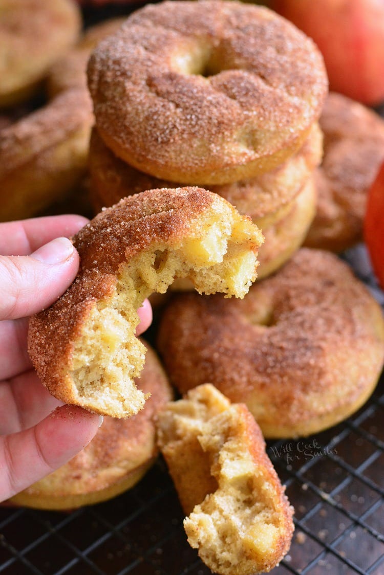 Apple Pie Baked Doughnuts with a piece of donut in my hand and a stack of apple doughnuts in the background on a cooling rack 