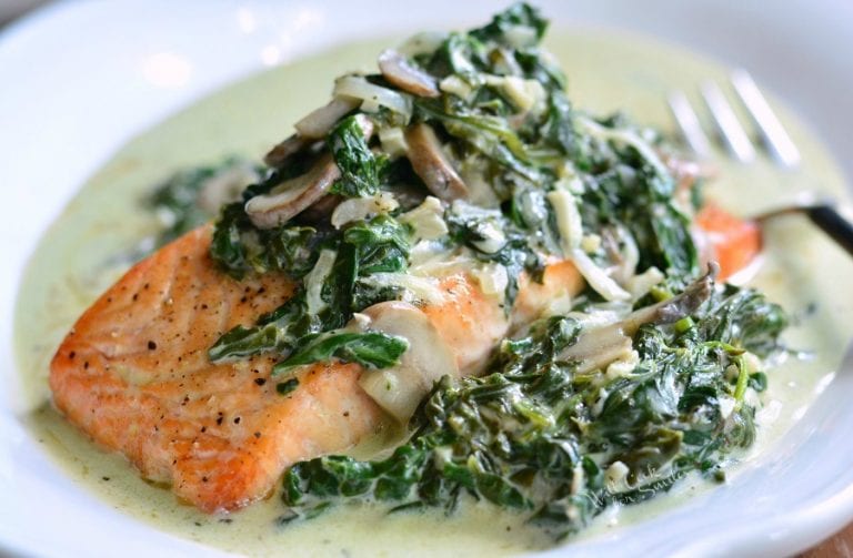 A plate of cooked salmon topped with creamy spinach and mushrooms, garnished with black pepper. The dish sits in a light green sauce, and a fork is visible in the background.