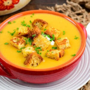 a red bowl full of bright orange butternut squash potato soup with croutons.