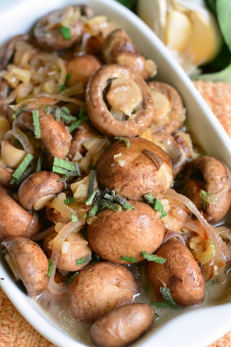 Mushrooms with Garlic and Sage in White Wine Sauce in a white baking dish with a spoon scooping some out on a cutting board with garlic in the background.