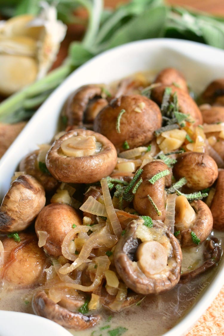 Mushrooms with Garlic and Sage in White Wine Sauce in a white baking dish on a cutting board with garlic in the background.