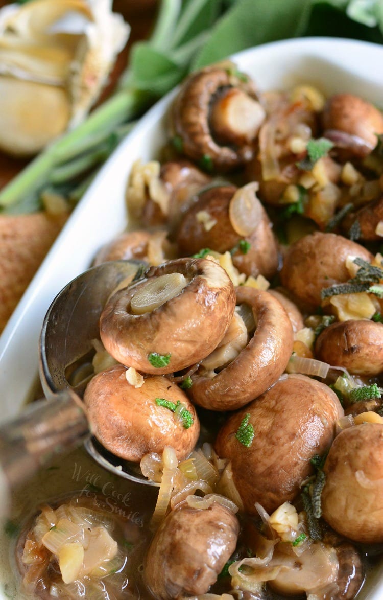 Mushrooms with Garlic and Sage in White Wine Sauce in a white baking dish with a spoon scooping some out on a cutting board with garlic in the background.