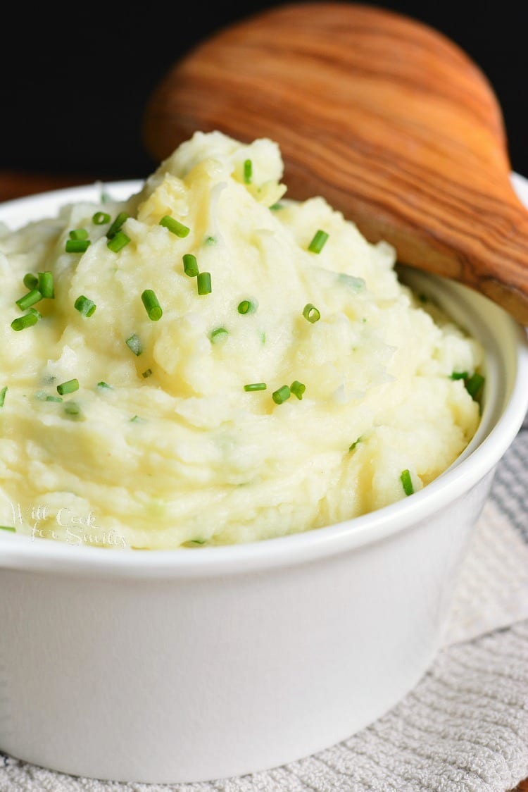 White Cheddar and Chive Creamy Mashed Potatoes in a bowl with chives on top and a wooden spoon 