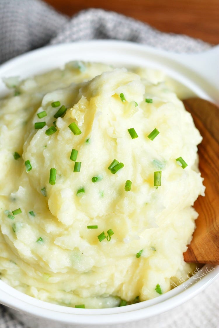 White Cheddar and Chive Creamy Mashed Potatoes in a serving bowl with chives on top and a wooden spoon 