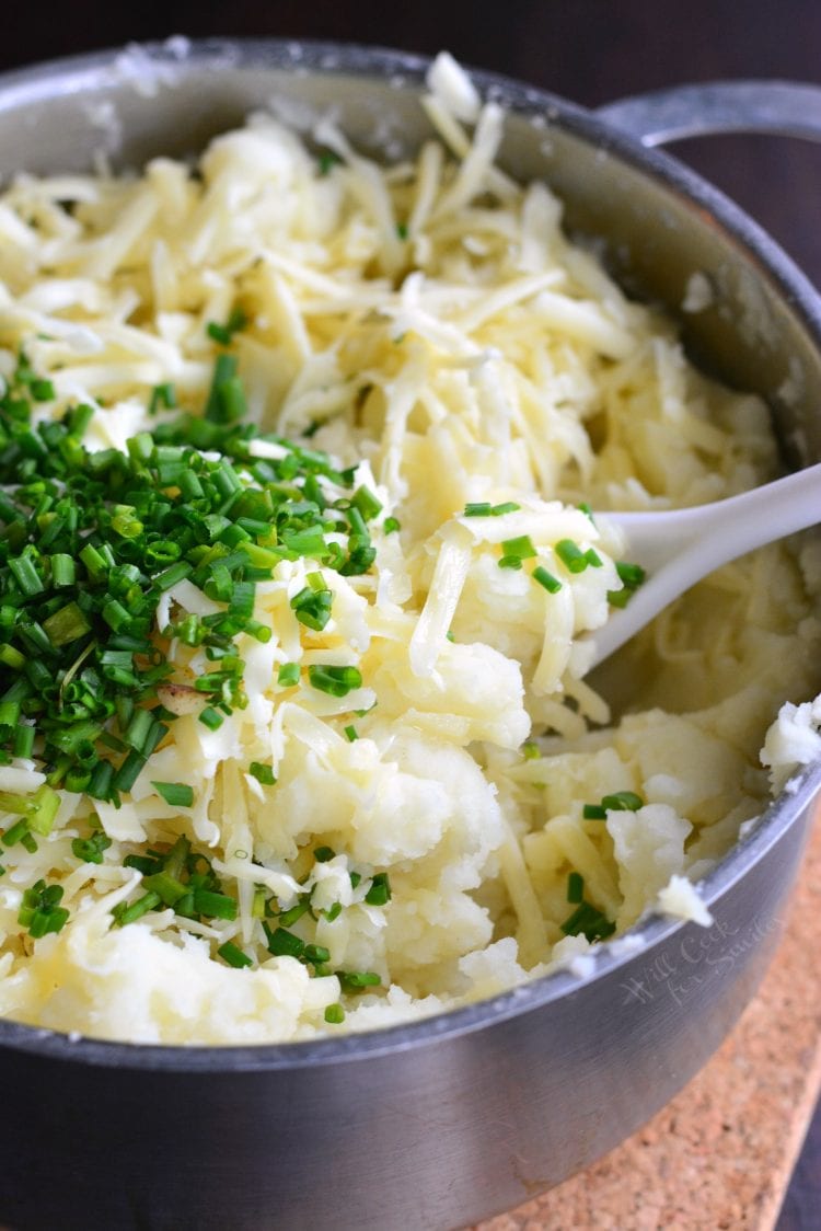 White Cheddar and Chive Creamy Mashed Potatoes in a pot with cheese and chives and a plastic spoon 