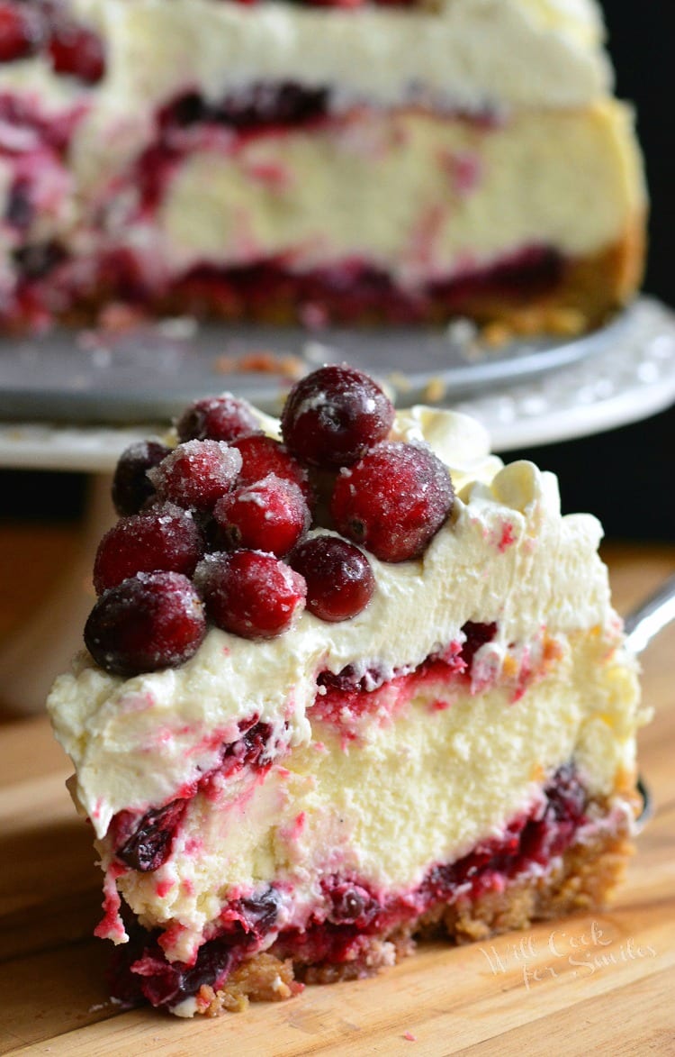 Christmas Cheesecake slice on a wood cutting board with cheesecake on stand in background 