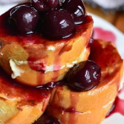 cherry cheesecake with cherries all over the top stacked on a white plate.