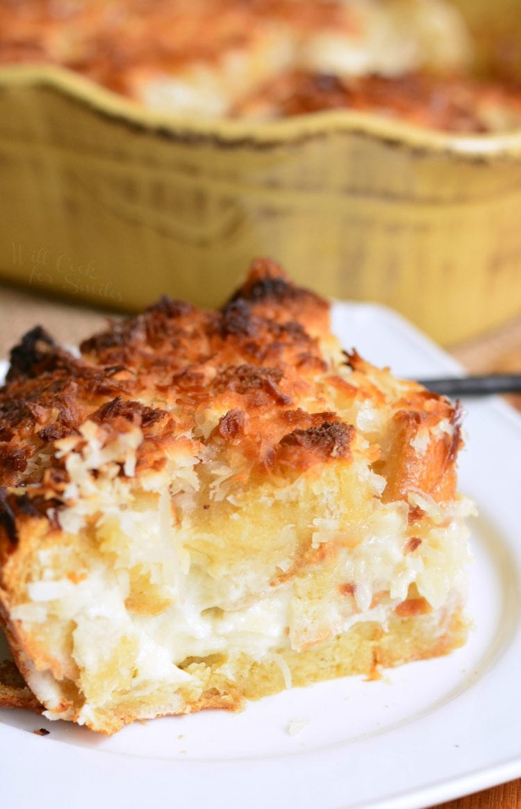 close up of coconut cheesecake bread putting on a white plate with a yellow casserole dish with the rest of the bread putting in the background.