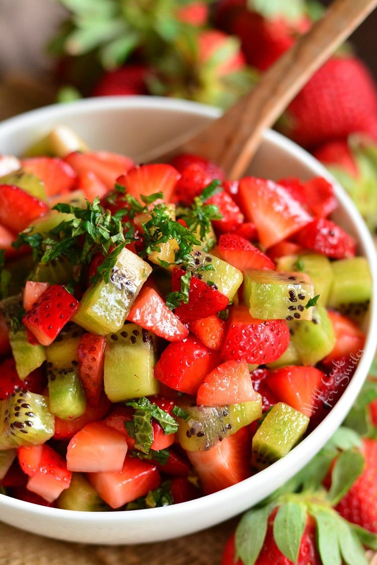 Fruit salad with kiwi and strawberries in a white bowl with a wooden spoon.