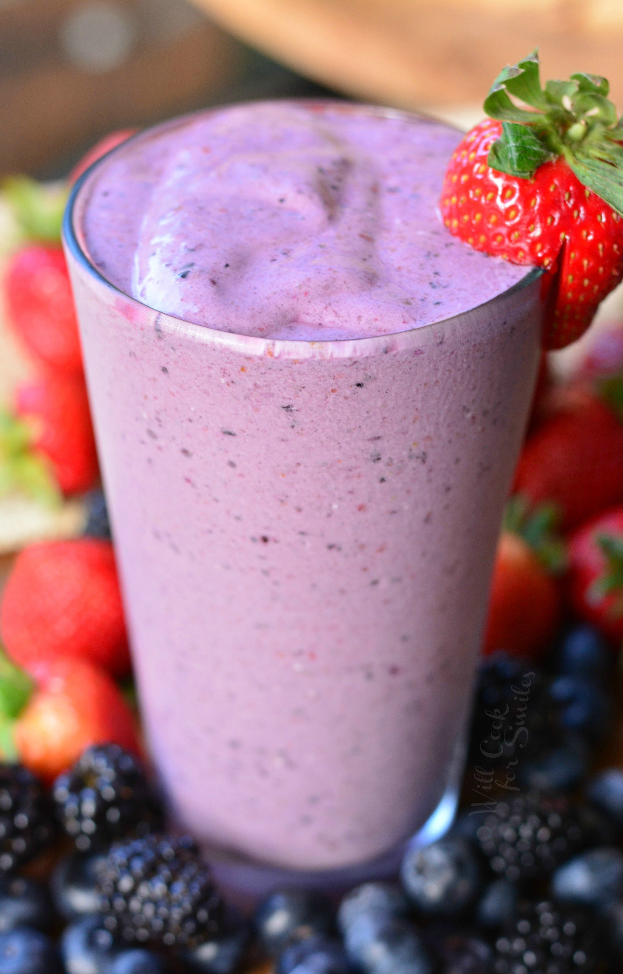 Berry Milkshake in a class with whip cream and strawberry to garnish on a wood table with blackberries, strawberries, and blueberries