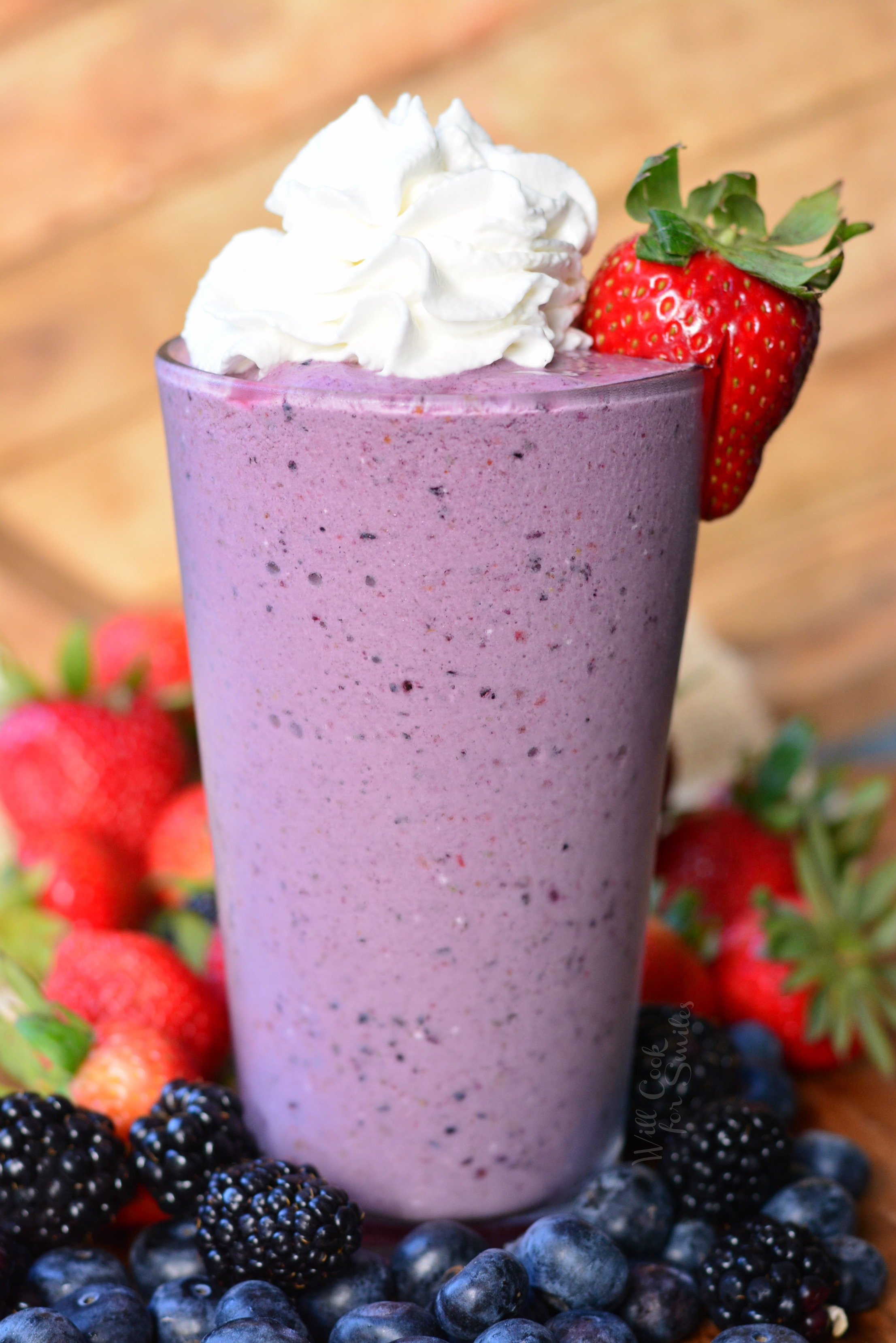 Berry Milkshake in a class with whip cream and strawberry to garnish on a wood table with blackberries, strawberries, and blueberries