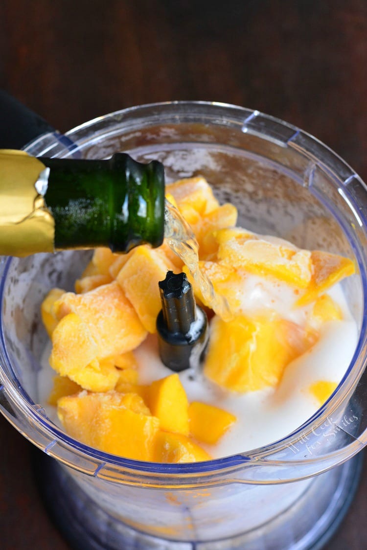 pouring champagne into a food processor with frozen mango.