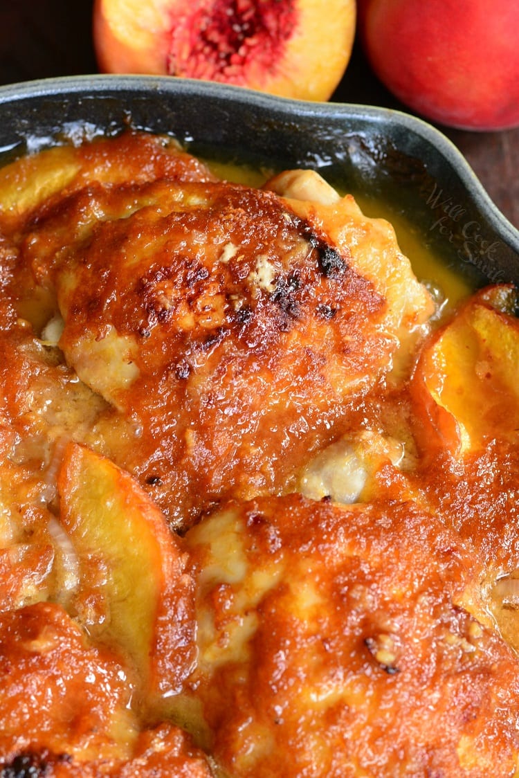 close up of two chicken thighs with sauce and peaches in green cast iron pan and peaches on the background